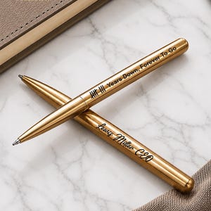 Op de afbeelding: Twee goudkleurige metalen balpennen met zwarte gravure. De ene pen bevat streepjes en de tekst "Years Down, Forever To Go", terwijl de andere pen is gegraveerd met de naam "Lewis Miller CEO" in een cursief lettertype.