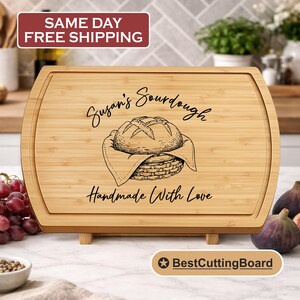 May include: A rectangular bamboo cutting board with rounded edges. The board features an engraved illustration of a loaf of bread in a basket, with the text "Susan's Sourdough" and "Handmade With Love". The board is supported by small wooden feet.