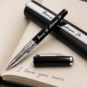 Puede incluir: Un bol&iacute;grafo negro y plateado con la inscripci&oacute;n "Thomas W. Anderson Jr." se muestra en un cuaderno abierto. El bol&iacute;grafo est&aacute; junto a su estuche, que tambi&eacute;n tiene el nombre grabado. El cuaderno dice "I love you more."
