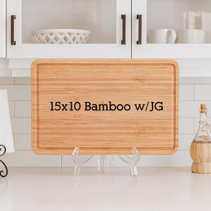 May include: A rectangular bamboo cutting board, 15 x 10 inches, with a juice groove. The text "15x10 Bamboo w/JG" is printed on the board. The board is displayed on a clear plastic stand.