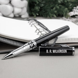 Puede incluir: Un bol&iacute;grafo rollerball negro y plateado con el texto "B. R. Wilkinson" en la tapa. El bol&iacute;grafo est&aacute; sobre una superficie gris junto a un cuaderno abierto. El bol&iacute;grafo tiene una banda decorativa plateada.