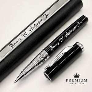 Puede incluir: Un bol&iacute;grafo negro y plateado con el nombre "Thomas W. Anderson Jr." grabado en el cuerpo y el estuche. El bol&iacute;grafo tiene una punta plateada y una tapa plateada y negra. El bol&iacute;grafo se muestra con su estuche, y las palabras "Premium Collection" est&aacute;n debajo.