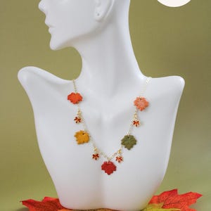 May include: A gold-tone chain necklace featuring multiple small, textured autumn leaf charms in shades of red, orange, yellow, and green. The necklace is displayed on a white mannequin bust, with the text Crafted LEVI by visible in the corner.