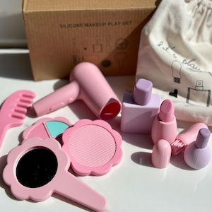 May include: A pink silicone makeup play set featuring a comb, hair dryer, mirrors, lipstick, nail polish, and a perfume bottle. The set includes a cardboard box and a drawstring bag. The box is printed with the text "SILICONE MAKEUP PLAY SET".