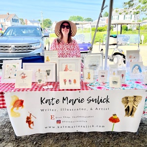 Op de afbeelding: Een vrouw staat achter een tafel met kunstprints en kaarten op een markt in de open lucht. De tafel is bedekt met een rood-wit geruite tafelkleed. De banner toont "Kate Marie Sulick, Writer, Illustrator, & Artist."
