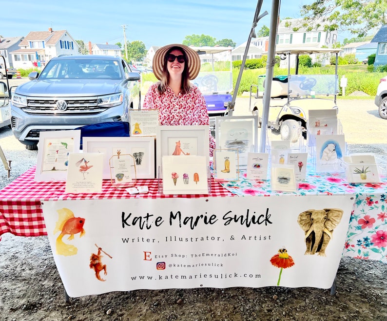 Op de afbeelding: Een vrouw staat achter een tafel met kunstprints en kaarten op een markt in de open lucht. De tafel is bedekt met een rood-wit geruite tafelkleed. De banner toont "Kate Marie Sulick, Writer, Illustrator, & Artist."