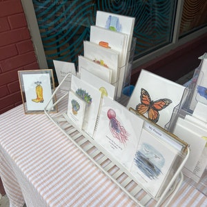 Op de afbeelding: Een display van kunstprints met aquarel illustraties. De prints worden weergegeven in een wit draadrek en een goud omrand display. De kunst omvat afbeeldingen van een vlinder, een kwal en een cowboylaars.