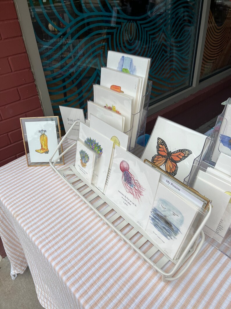Op de afbeelding: Een display van kunstprints met aquarel illustraties. De prints worden weergegeven in een wit draadrek en een goud omrand display. De kunst omvat afbeeldingen van een vlinder, een kwal en een cowboylaars.