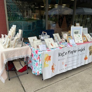 Op de afbeelding: Een kunstkraam in de openlucht met ingelijste illustraties, prints en kaarten. De tafel is bedekt met een bloemenkleed en een gestreepte tafelkleed met franjes. De banner luidt "Kate Marie Sulick, Writer, Illustrator, & Artist."