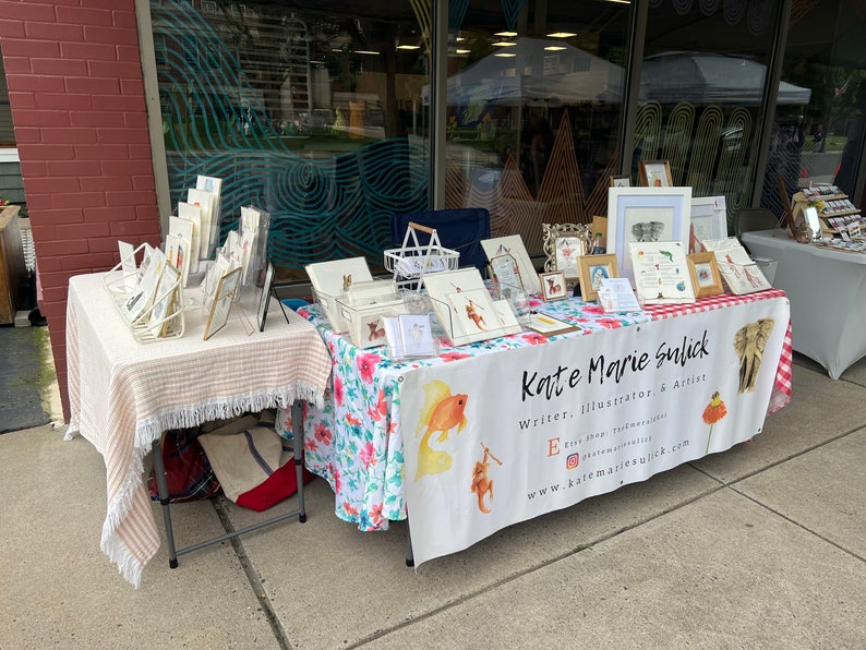 Op de afbeelding: Een kunstkraam in de openlucht met ingelijste illustraties, prints en kaarten. De tafel is bedekt met een bloemenkleed en een gestreepte tafelkleed met franjes. De banner luidt "Kate Marie Sulick, Writer, Illustrator, & Artist."