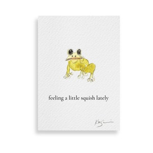 Op de afbeelding: Een aquarelprint met een gele kikker met grote ogen en een klein voorwerp in zijn mond. De tekst "feeling a little squish lately" staat onder de kikker. Het kunstwerk is rechtsonder gesigneerd.