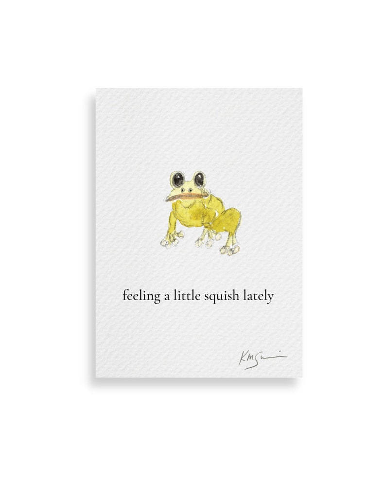 Op de afbeelding: Een aquarelprint met een gele kikker met grote ogen en een klein voorwerp in zijn mond. De tekst "feeling a little squish lately" staat onder de kikker. Het kunstwerk is rechtsonder gesigneerd.