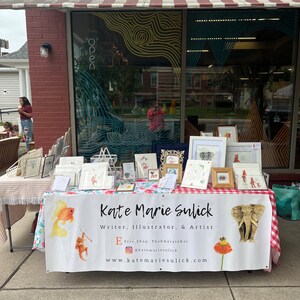 Op de afbeelding: Een marktkraam met kunstwerken en kaarten. Een banner leest "Kate Marie Sulick, Writer, Illustrator, & Artist." Een gouden olifant sculptuur is te zien. De luifel erboven is rood en wit gestreept.