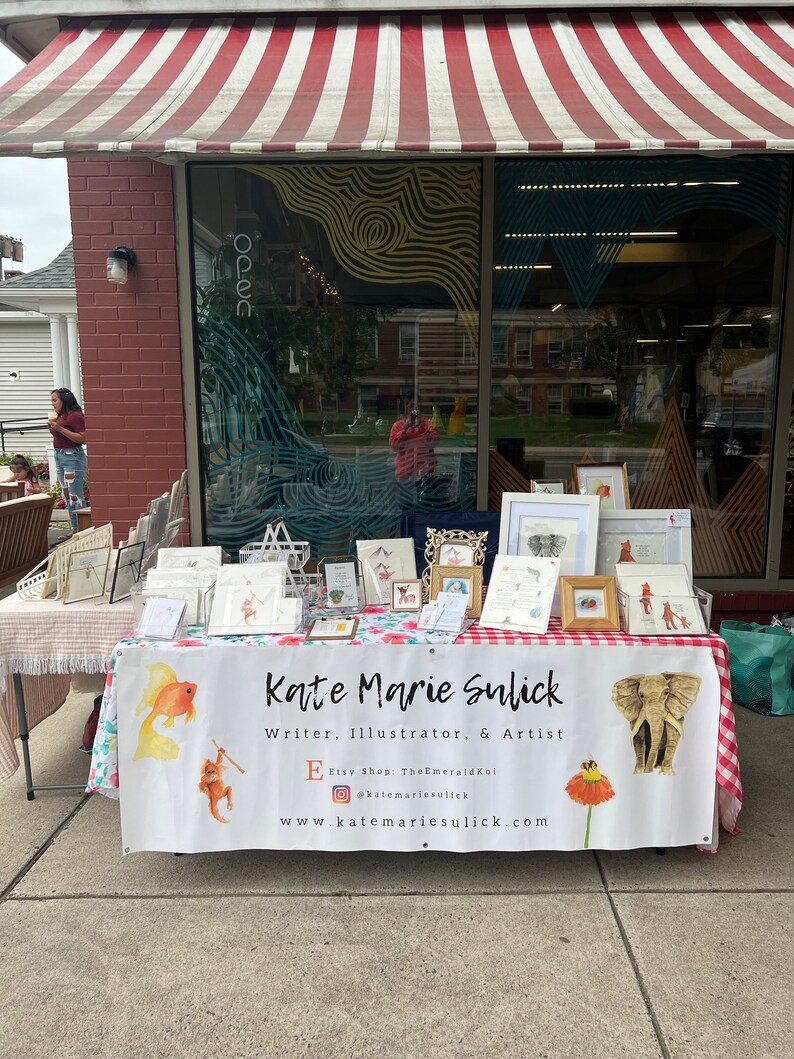 Op de afbeelding: Een marktkraam met kunstwerken en kaarten. Een banner leest "Kate Marie Sulick, Writer, Illustrator, & Artist." Een gouden olifant sculptuur is te zien. De luifel erboven is rood en wit gestreept.