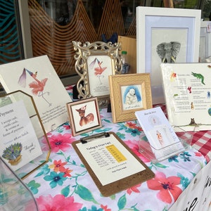 Op de afbeelding: Een marktkraam met ingelijste kunstafdrukken en kaarten. De kunstwerken omvatten aquarel schilderijen van dieren, zoals een hert, een flamingo en een olifant. Een prijslijst en kunstenaarsinformatie zijn ook zichtbaar.