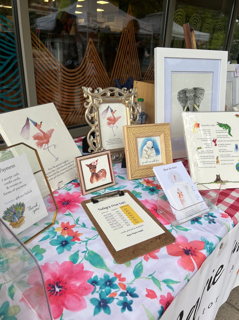 Op de afbeelding: Een marktkraam met ingelijste kunstafdrukken en kaarten. De kunstwerken omvatten aquarel schilderijen van dieren, zoals een hert, een flamingo en een olifant. Een prijslijst en kunstenaarsinformatie zijn ook zichtbaar.