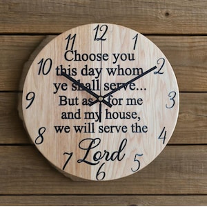 May include: A round wooden clock with black numerals and hands. The clock face features the quote "Choose you this day whom ye shall serve... But as for me and my house, we will serve the Lord." The clock is mounted on a wooden background.