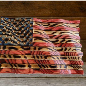 May include: A wooden American flag with a wavy design. The flag features the blue field with stars and red and white stripes. The wood has a natural grain and is stained with red, white, and blue colors.