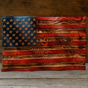 May include: A wooden American flag with a distressed finish. The flag features a blue field with carved stars and red and white stripes. The wood has a burnt, textured appearance, giving it a rustic look.