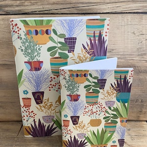 May include: Two notebooks with a cream background and a colourful pattern of potted plants. The pattern includes various plants, pots, and colours. The notebooks are different sizes.