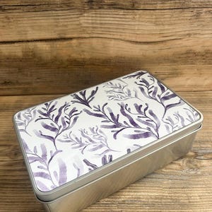 May include: A rectangular metal tin with a lid decorated with a watercolour design of purple seaweed on a white background. The tin has a silver finish and is on a wooden surface.