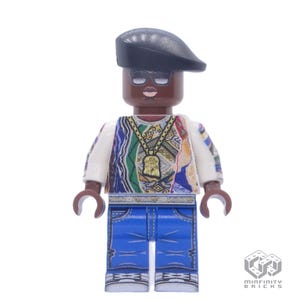 Op de afbeelding: Een Lego-minifiguur van een persoon met een zwarte baret, zonnebril en gouden ketting. De figuur draagt een kleurrijke trui, een wit overhemd en een blauwe spijkerbroek. De figuur heeft een bruine huid en staat rechtop.