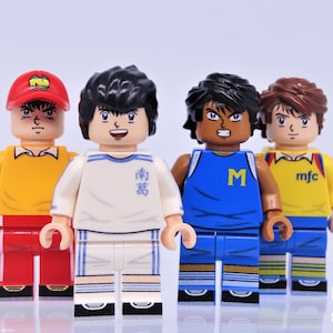 May include: Four minifigure toys of soccer players wearing different uniforms. The players are wearing red, white, blue, and yellow jerseys. The players have different hairstyles and facial expressions. The minifigures are standing on a white surface.