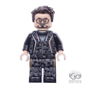 May include: A Lego minifigure of Iron Man wearing a black and grey suit with a silver arc reactor on his chest. The minifigure has a brown hairpiece and a printed face with a beard and glasses.