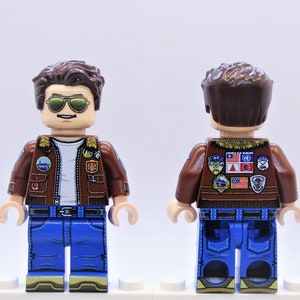 Puede incluir: Una chaqueta de cuero marr&oacute;n con cuello de piel y varios parches, usada por una minifigura de Lego con cabello marr&oacute;n, gafas de sol y jeans azules.