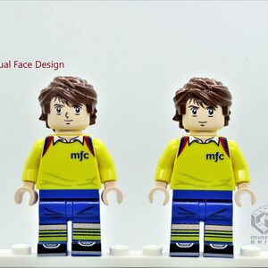 May include: Dual face design Lego minifigure with brown hair, yellow shirt with "mfc" printed on the front, blue pants, and yellow and white striped socks. The minifigure is standing on a white surface.