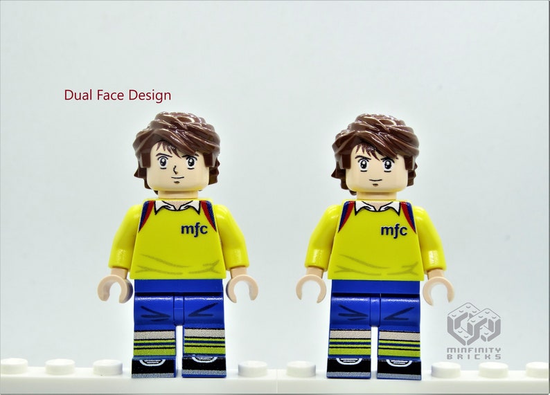 May include: Dual face design Lego minifigure with brown hair, yellow shirt with "mfc" printed on the front, blue pants, and yellow and white striped socks. The minifigure is standing on a white surface.