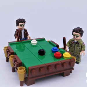Puede incluir: Dos figuras de Lego jugando al billar en una mesa de billar de fieltro verde con patas de madera marr&oacute;n. La mesa tiene seis bolsillos y las figuras sostienen tacos de billar.