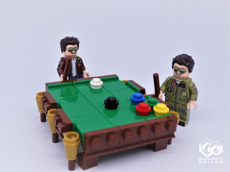 Puede incluir: Dos figuras de Lego jugando al billar en una mesa de billar de fieltro verde con patas de madera marr&oacute;n. La mesa tiene seis bolsillos y las figuras sostienen tacos de billar.