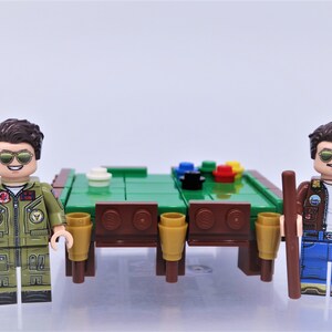 Puede incluir: Dos personajes de minifigura con cabello casta&ntilde;o y gafas de sol, vistiendo chaquetas verdes y azules, sosteniendo tacos de billar marrones, de pie a ambos lados de una mesa de billar de fieltro verde con lados de madera marr&oacute;n y seis portavasos marrones y dorados.