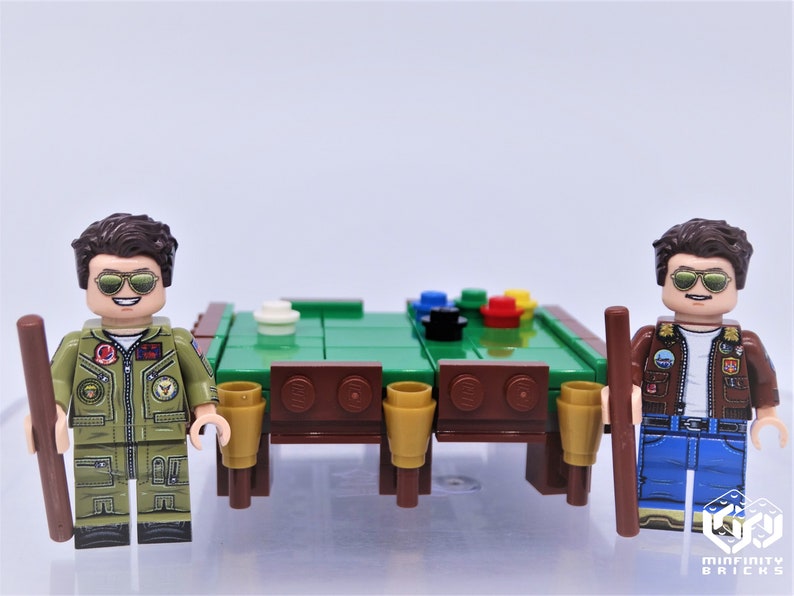 Puede incluir: Dos personajes de minifigura con cabello casta&ntilde;o y gafas de sol, vistiendo chaquetas verdes y azules, sosteniendo tacos de billar marrones, de pie a ambos lados de una mesa de billar de fieltro verde con lados de madera marr&oacute;n y seis portavasos marrones y dorados.