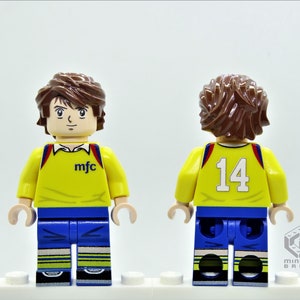 May include: A yellow and blue Lego minifigure wearing a soccer jersey with the text "mfc" on the front and the number "14" on the back.