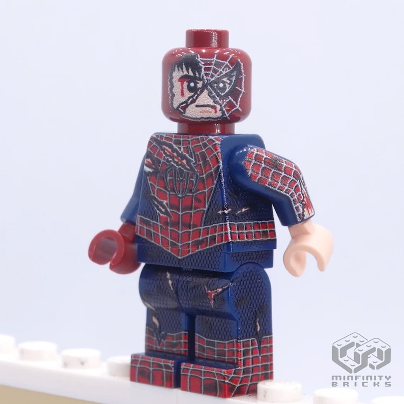 May include: A custom Lego minifigure of Spider-Man in a distressed state. The figure features a red and blue costume with a web design, and a damaged mask revealing a portion of the face. The figure is standing on a white surface.