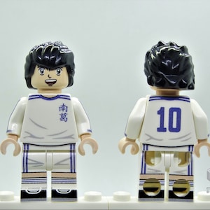 May include: A white and blue Lego minifigure of a soccer player wearing a white jersey with blue accents and the number 10 on the back. The jersey has the Japanese characters "南葛" on the front. The minifigure has black hair and a smiling face.