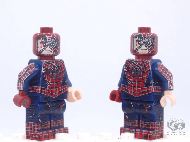 May include: Two Lego Spider-Man minifigures. Each figure has a dark red head with a distressed mask design and a blue and red suit. The suit features a spiderweb pattern and the word "AM" on the back. The figures are standing against a white background.