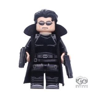 May include: A black Lego minifigure of a man wearing a black trench coat, black sunglasses, and holding two black guns. The minifigure has a black belt and black trousers.