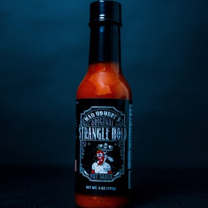 May include: A glass bottle of Mad Hombre's Original Strangle Hold hot sauce. The bottle is black with a white label and a cartoon illustration of a man with a red face and a sombrero. The label reads "Mad Hombre's Original Strangle Hold Hot Sauce" and "Net Wt. 5 oz (141g)".