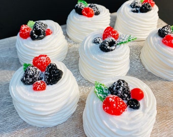 May include: Six mini cakes with white frosting and a variety of fresh berries on top. The cakes are arranged on a white wooden surface.