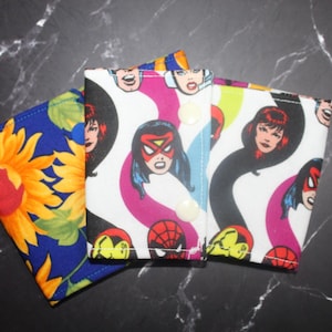 May include: Three fabric items with colourful patterns. One features a sunflower design on a blue background. The other two have a comic book theme with superhero faces and logos on a white background with pink, yellow, and black accents.