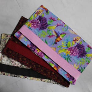 May include: A selection of fabric-covered journals and notebooks. The top notebook displays a colourful floral and hummingbird design with a pink elastic band. Other notebooks have burgundy, black, and patterned covers.