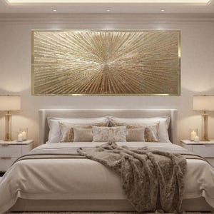 Pode incluir: Uma cena de quarto com uma grande obra de arte abstrata dourada acima de uma cama. A obra de arte tem um design radial. A cama é feita com roupa de cama e almofadas de cor neutra, com uma manta castanha. Duas mesas de cabeceira com lâmpadas completam a cena.