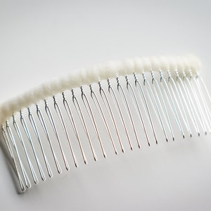 May include: A silver hair comb with a white, fuzzy covering on the top. The comb has a curved shape and is designed for holding hair in place.