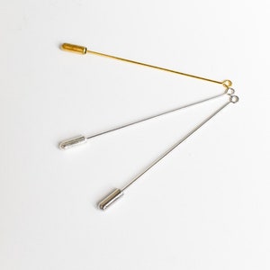 May include: Three stick pins in gold and silver tones. The gold pin has a cylindrical head, while the silver pins have rounded heads. The pins are arranged on a white surface, showcasing their slender design and metallic sheen.