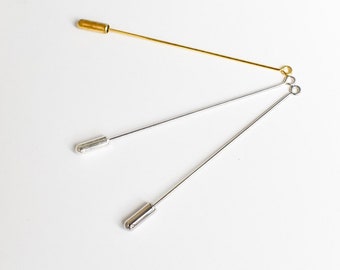 May include: Three stick pins in gold and silver tones. The gold pin has a cylindrical head, while the silver pins have rounded heads. The pins are arranged on a white surface, showcasing their slender design and metallic sheen.