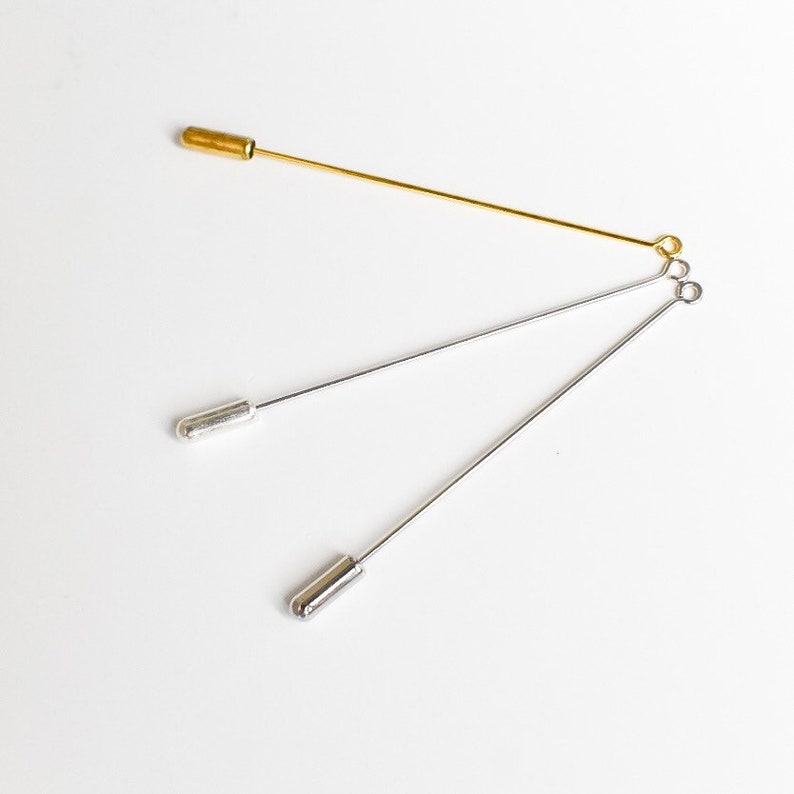 May include: Three stick pins in gold and silver tones. The gold pin has a cylindrical head, while the silver pins have rounded heads. The pins are arranged on a white surface, showcasing their slender design and metallic sheen.
