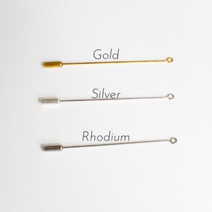 May include: Three metal lapel pins in gold, silver, and rhodium. Each pin has a straight, slender body with a rounded end and a small loop at the opposite end. The word "Gold", "Silver", and "Rhodium" are written above each pin.